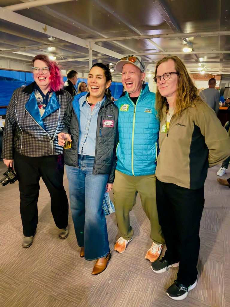 A group of four adults stands closely together indoors, smiling and laughing as they pose for a photo. From left to right: the first person has bright pink hair, glasses, and wears a dark, patterned jacket over black pants, holding a camera in their right hand. The second person has dark hair pulled back and wears a denim shirt under a navy jacket with flared blue jeans and brown boots, holding a drink. The third person wears a light gray cap, reflective sunglasses on their head, a bright teal puffer jacket, khaki pants, and orange sneakers. The fourth person has long, light brown hair and glasses, wearing a tan and black zip-up jacket with black pants and sneakers. Behind them, other people mingle in a well-lit, covered space with white beams, wood paneling, and a large window showing a view of water and distant land outside. The group appears happy and casual, enjoying a social event.